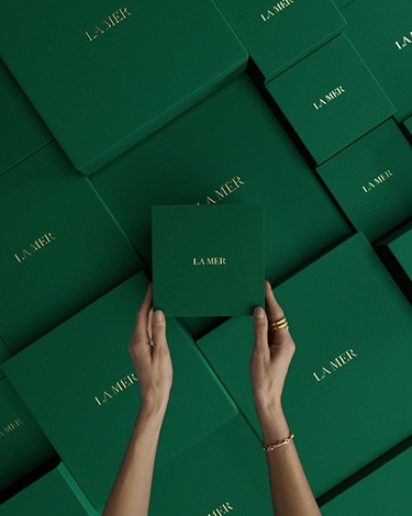 Hands with white gloves placing lid on green giftbox, then tying a gold la mer ribbon into a bow, cutting ends of ribbon.